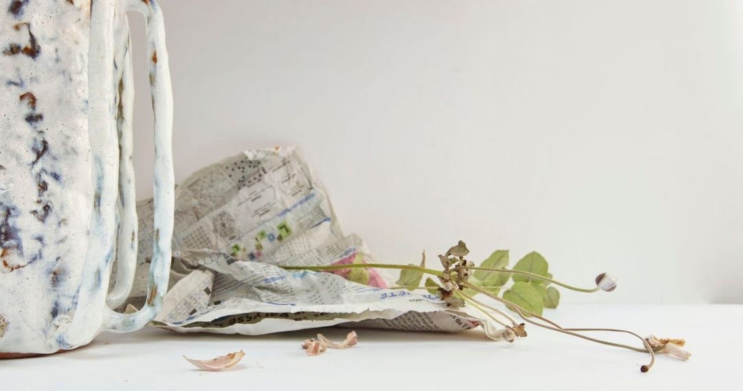 Still life shot featuring a glazed ceramic mug alongside dried flowers and foliage, set on a white surface with newspaper.
