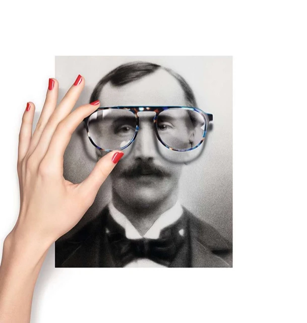 An eyeglasses frame is held up to an old portrait of a gentleman with a mustache, red nail polish on the manicured fingers holding the glasses.