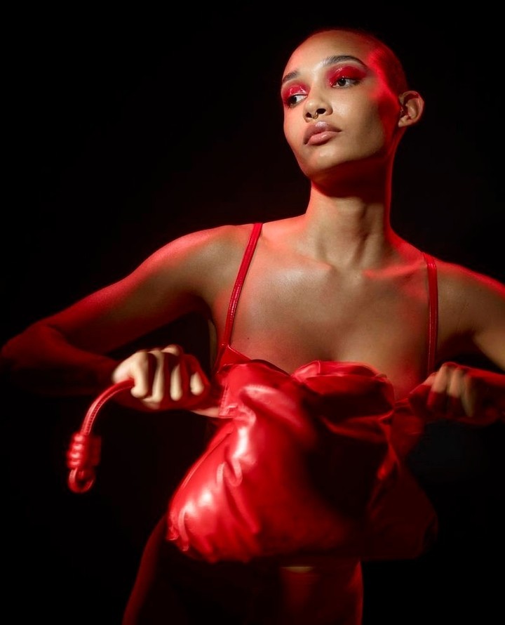 Close-up of a bald woman with red eyeshadow holding a red knotted bag in a dark studio setting with dramatic lighting.