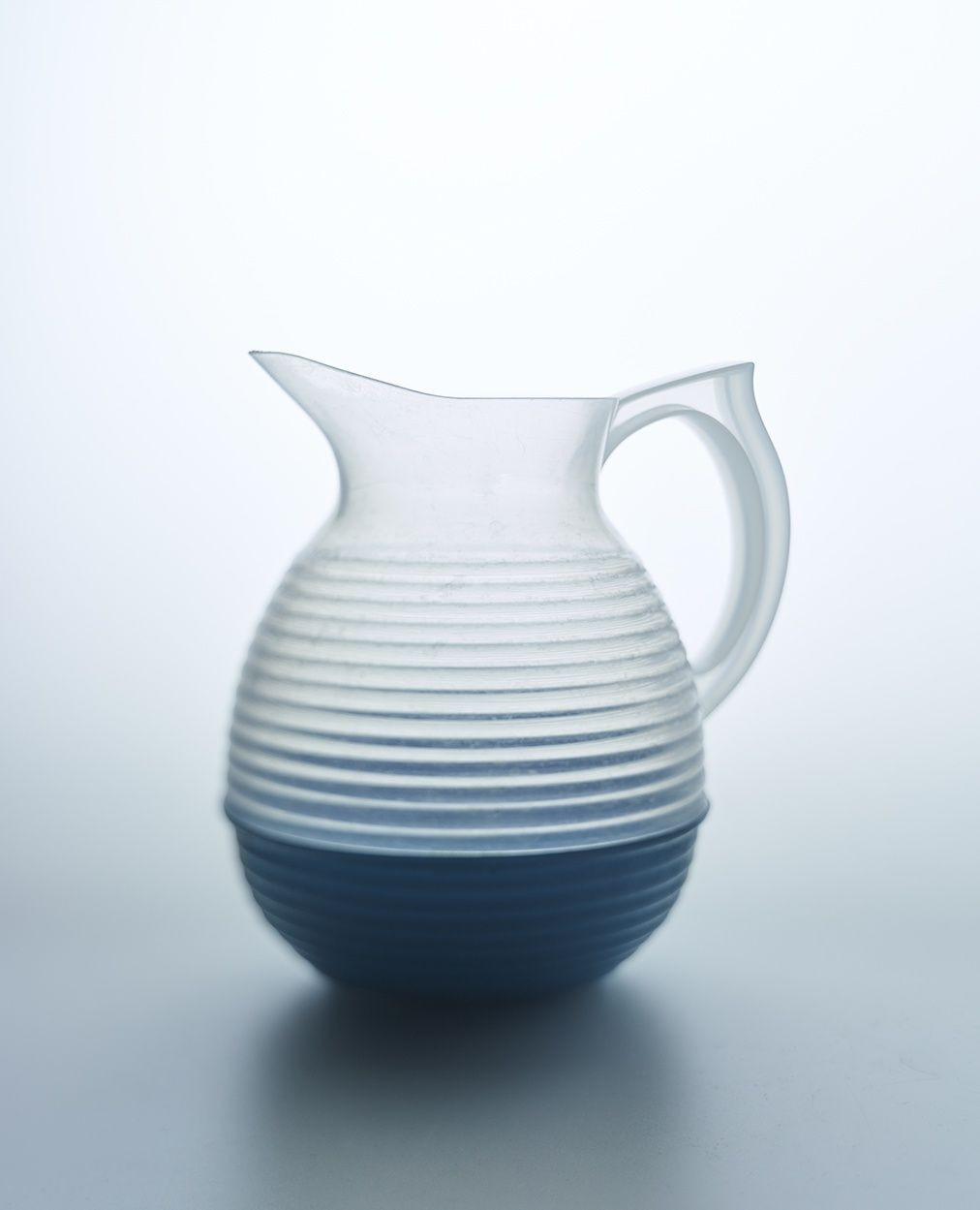 Studio shot of a clear glass pitcher with blue base and grooved design, set against a bright, neutral backdrop showcasing minimalist design.