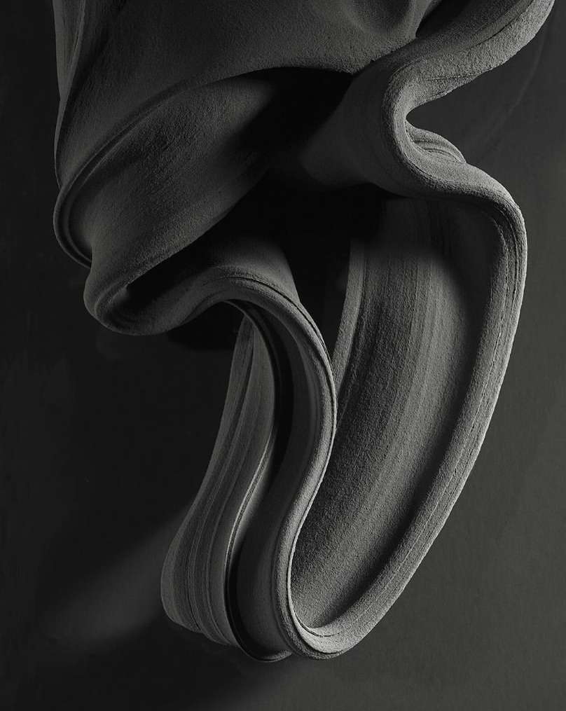 Abstract gray fabric drapes and swirls against a dark gray backdrop, creating a textured, monochrome artistic composition.