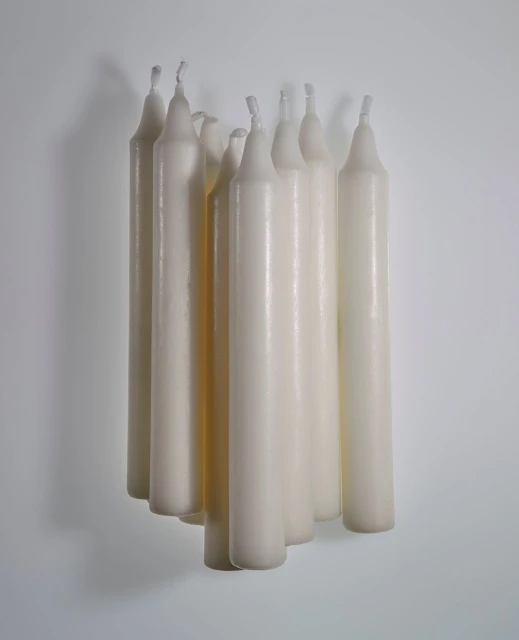 Elevated view of a collection of slender, ivory-colored taper candles clustered in a staggered arrangement against a soft gray backdrop.