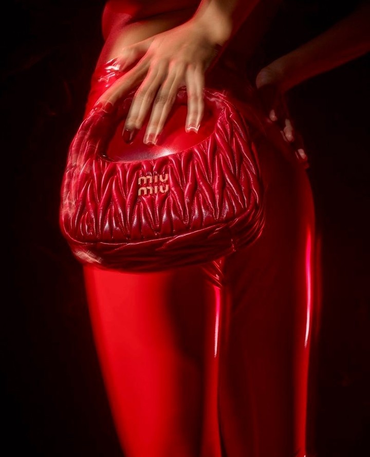 Close-up of a fashion model holding a red Miu Miu handbag while posing indoors in matching red pants and top, under a dark moody light.