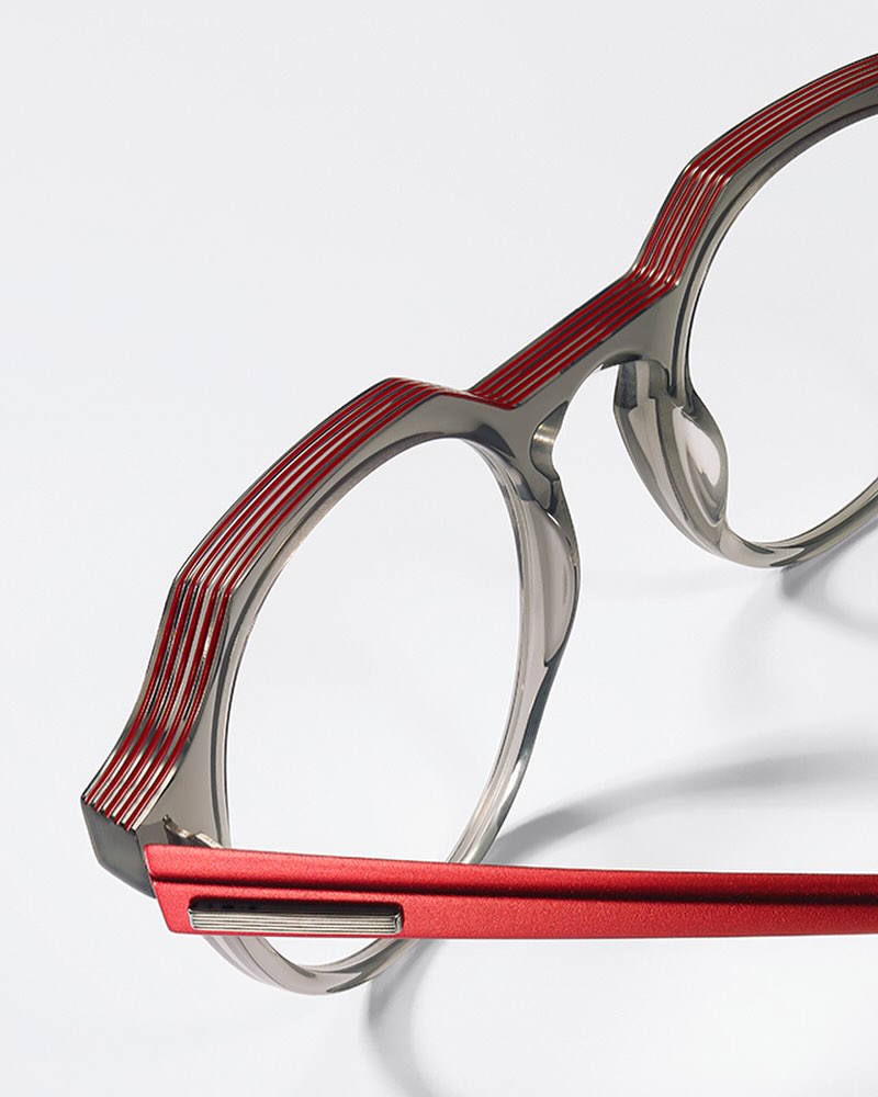 Close-up of round eyeglasses with gray rims, red detailing, and shadow on a white background; eye-wear fashion product shot.