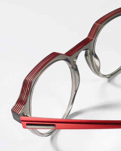 Close-up of round eyeglasses with gray rims, red detailing, and shadow on a white background; eye-wear fashion product shot.