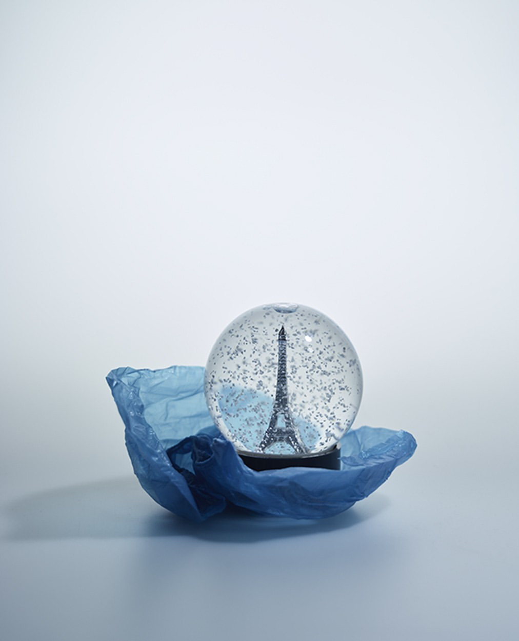 Snow globe with the Eiffel Tower inside, sitting on a blue plastic bag against a light blue background in a studio setting.