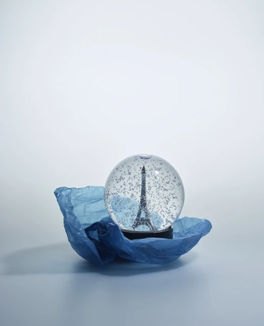 Snow globe with the Eiffel Tower inside, sitting on a blue plastic bag against a light blue background in a studio setting.