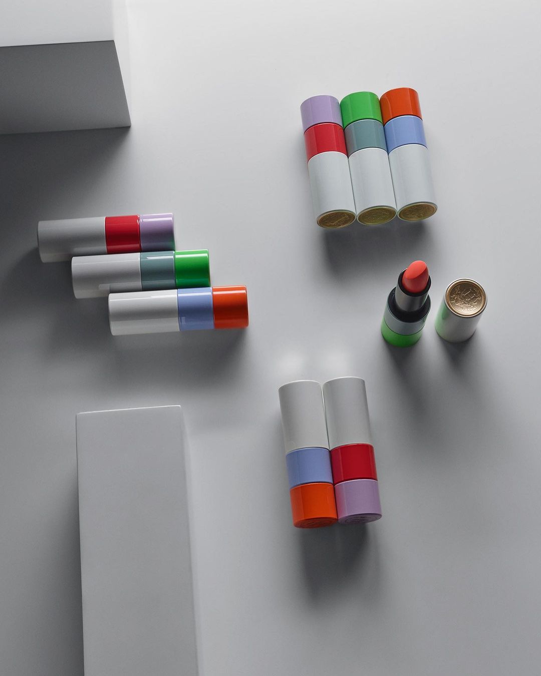 Product shot of stacked and arranged lipstick tubes in a variety of colors and finishes on a white surface with architectural elements.