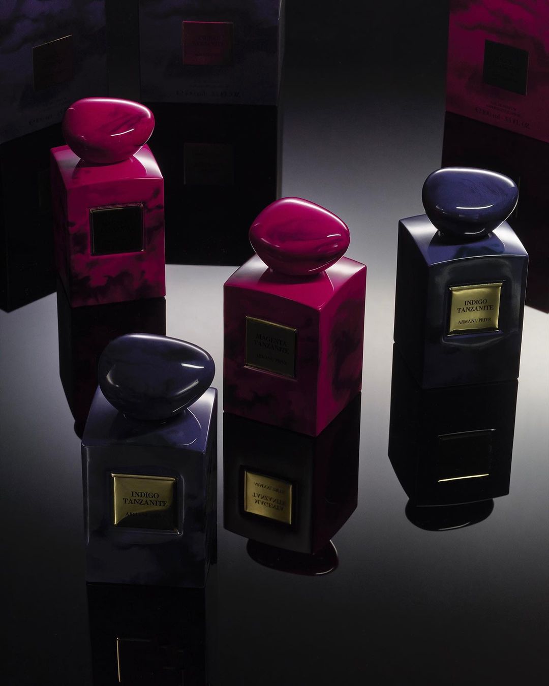 Four Giorgio Armani Indigo Tanzanite perfume bottles with black and magenta colors on a glossy black surface reflecting studio lighting.