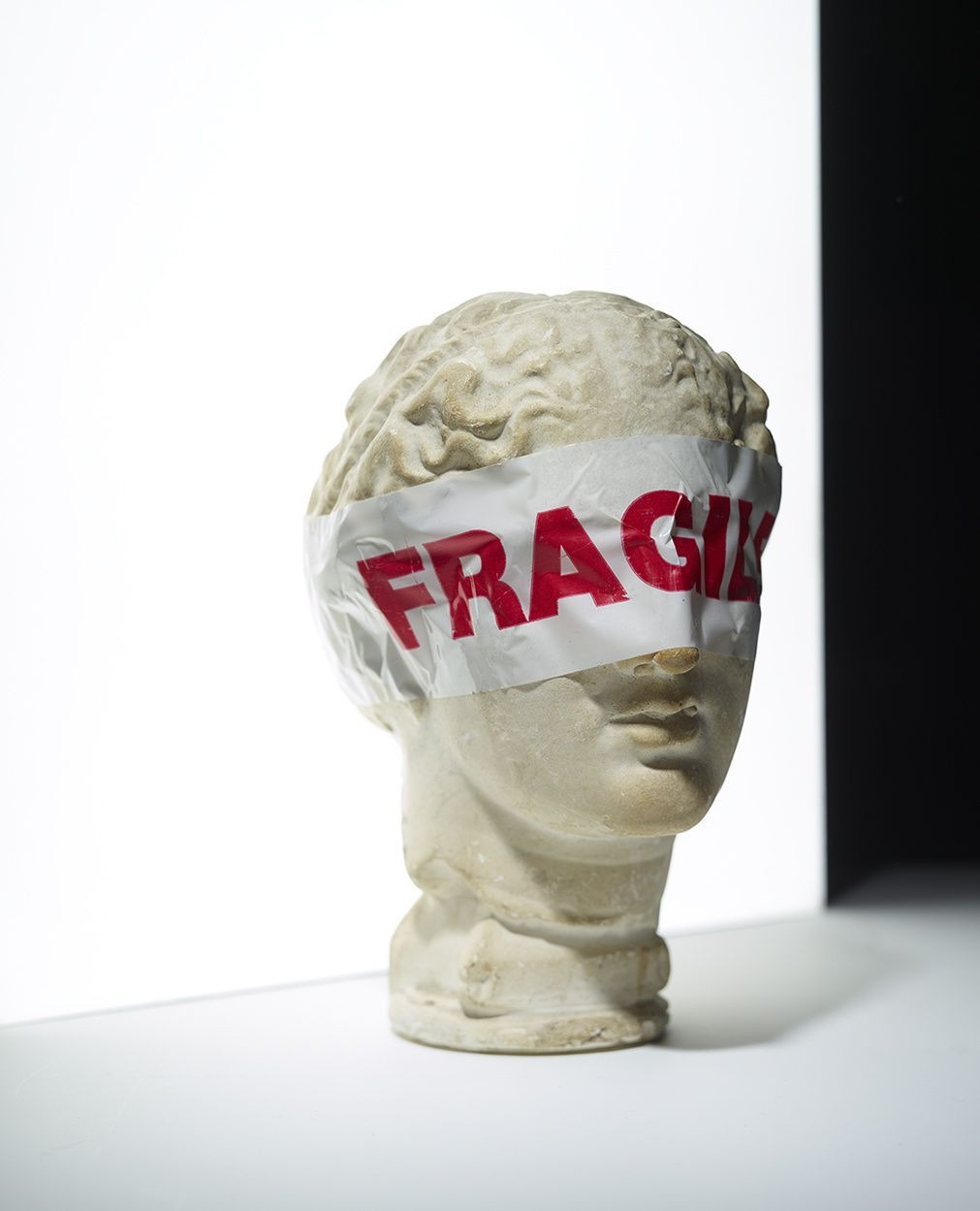Still life studio shot of a bust sculpture with a fragile label covering the eyes, against a white and black background.