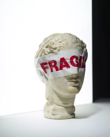 Still life studio shot of a bust sculpture with a fragile label covering the eyes, against a white and black background.