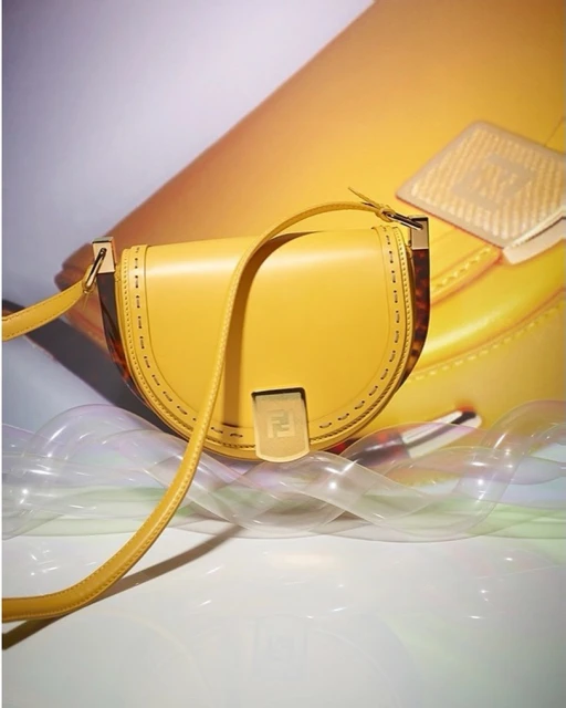 Yellow leather Fendi handbag on a white surface with iridescent plastic and a blurred bag in the background, studio product shot.
