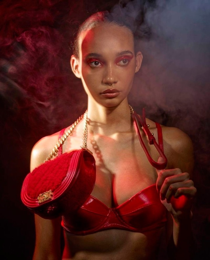 Woman poses in studio wearing red bra and gold chain with a red purse. Red eye makeup, smoke in background.