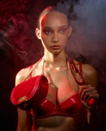 Model in red bra wearing a red crossbody bag with gold chain strap against smoky red and dark background