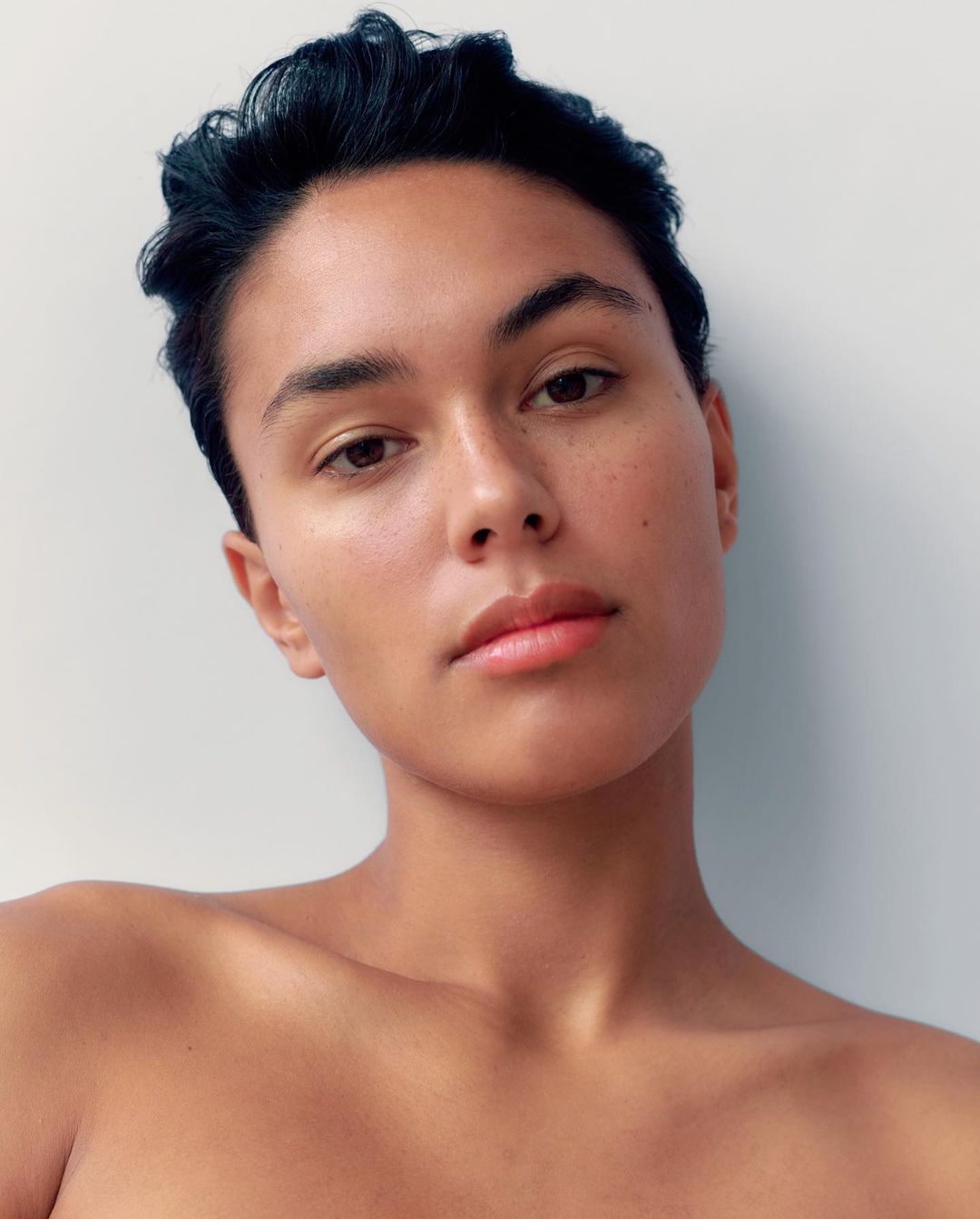 Close-up portrait of a young adult with short, dark hair against a simple white background, focusing on natural skin texture.