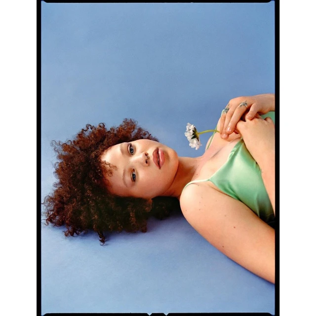 Portrait of a brunette woman lying on a blue surface, holding a daisy. Studio lighting highlights her features.