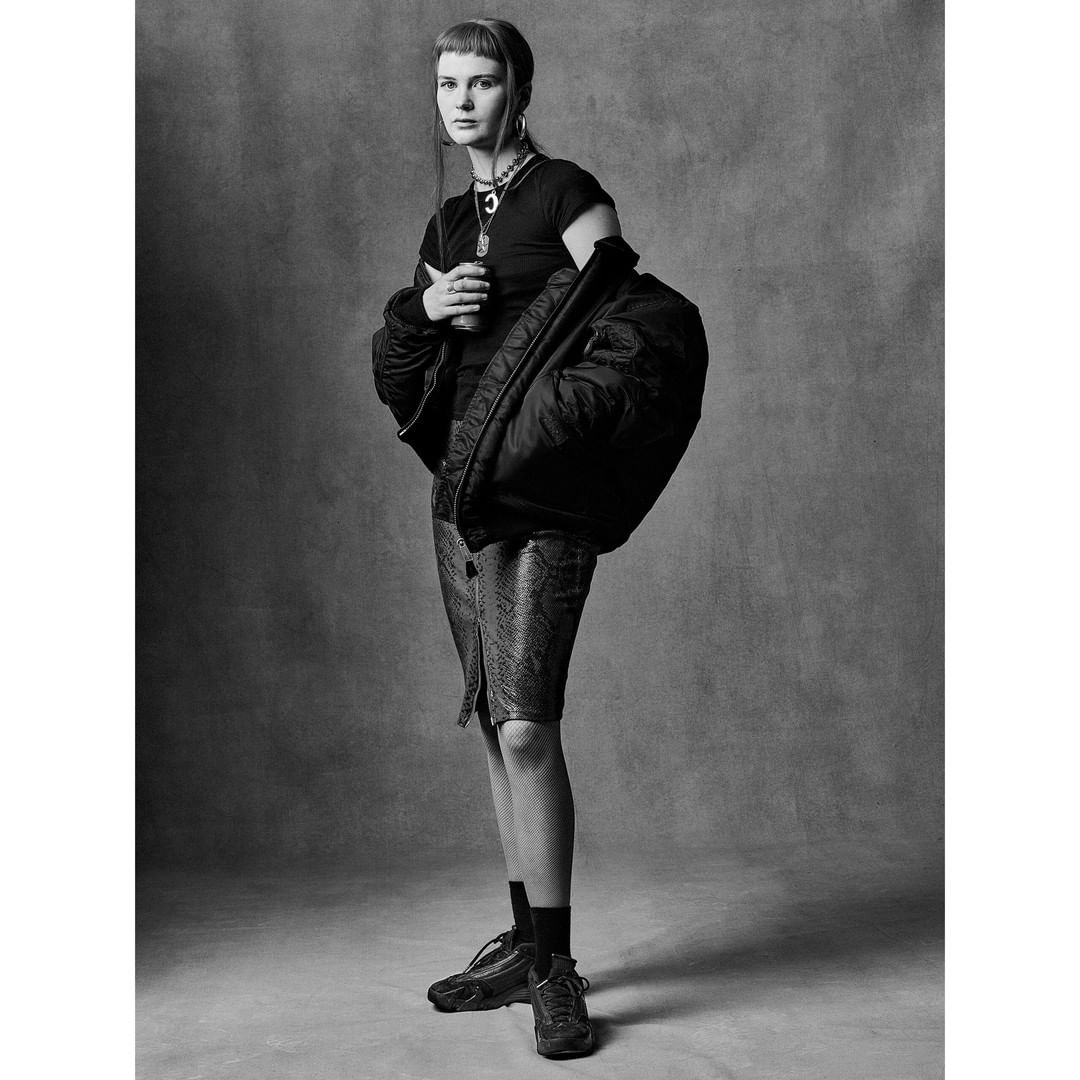 Monochrome studio portrait of an adult woman wearing a dark t-shirt, patterned skirt, and bomber jacket draped on her shoulders.