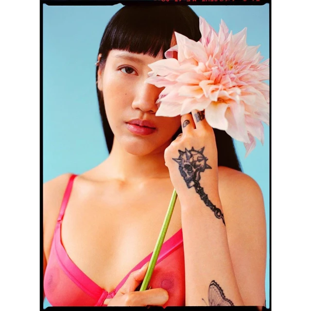 Close-up portrait of a woman with bangs holding a large pink flower, wearing a pink bra, and featuring arm tattoos against a blue background.