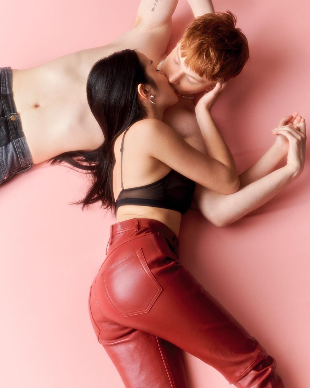 An intimate studio shot features a couple kissing while lying down against a pink backdrop, with a focus on their relaxed pose and clothing.