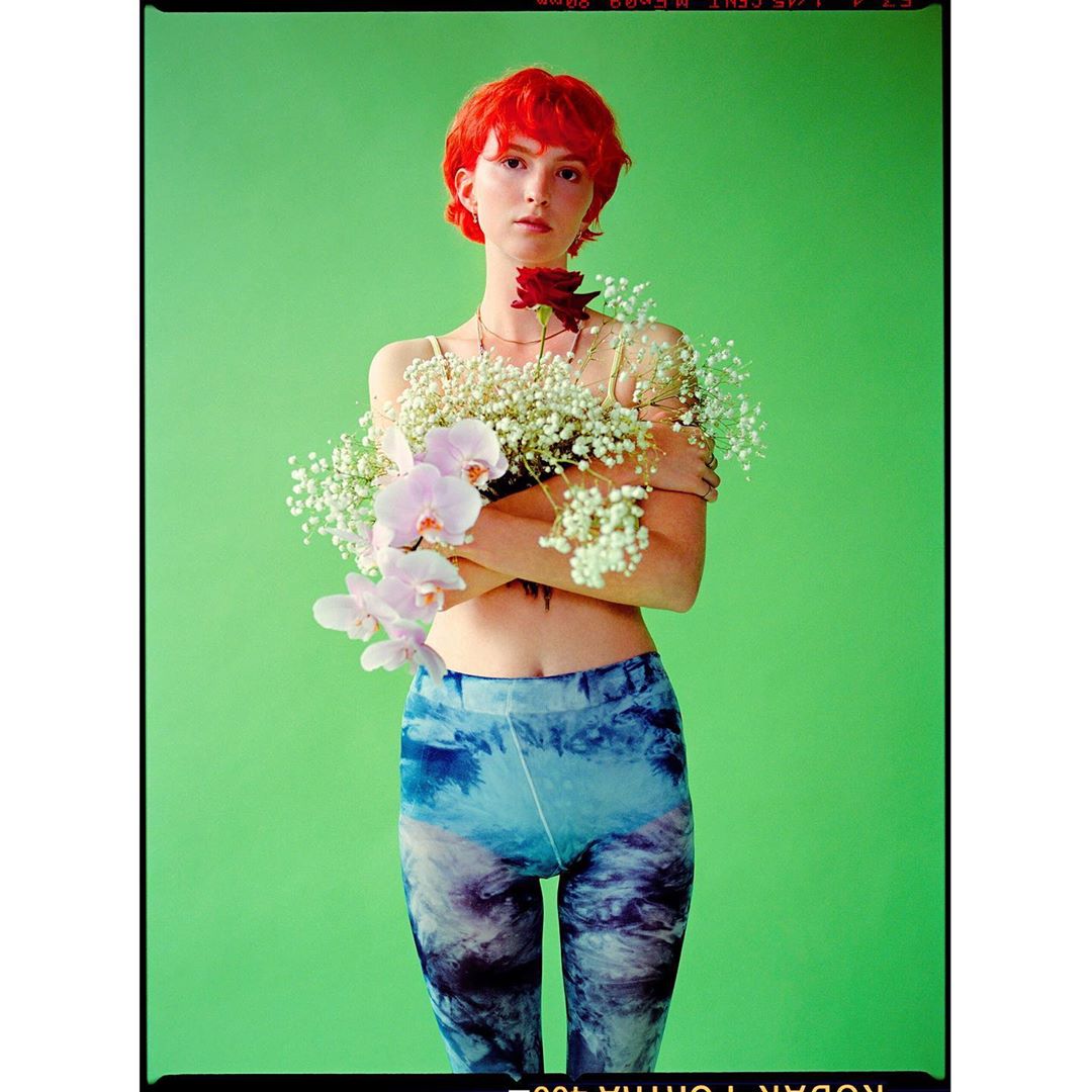 A young woman with red hair poses with a bouquet of white flowers and a red rose against a bright green backdrop in a studio portrait.