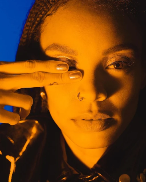 Close-up portrait of a woman with braided hair, nose ring, and hand partially covering one eye, illuminated by warm yellow lighting.