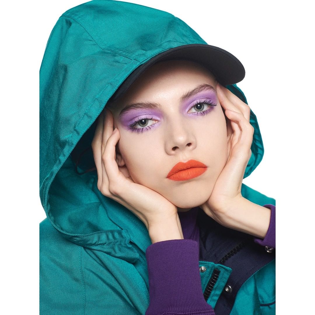 A light-skinned adult female model wears bright, contrasting makeup and a hooded teal jacket in a studio shot, showcasing beauty and fashion.