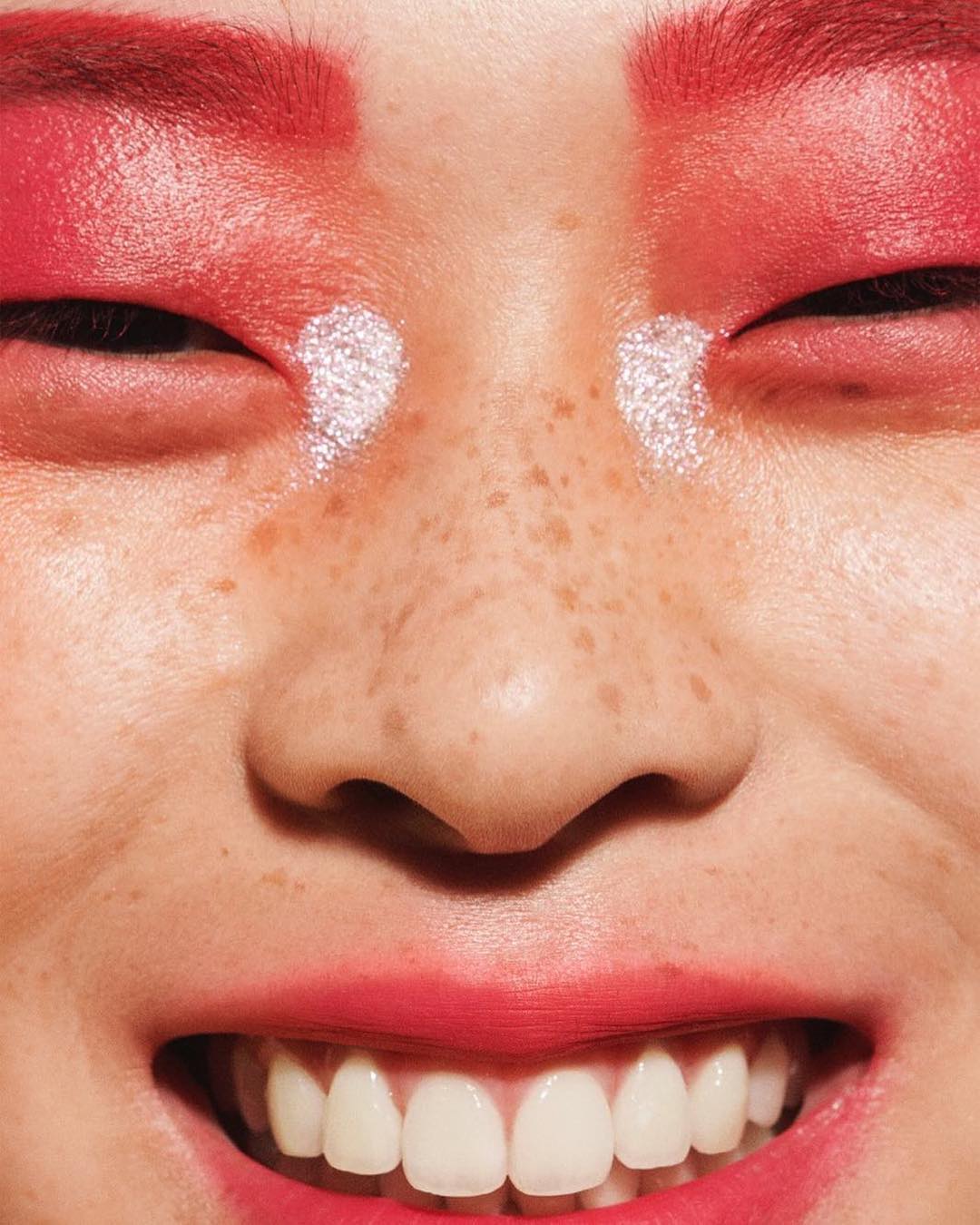 Close-up of a smiling adult female face with red makeup on eyebrows and eyelids, and white makeup under the eyes. Clear skin and white teeth.