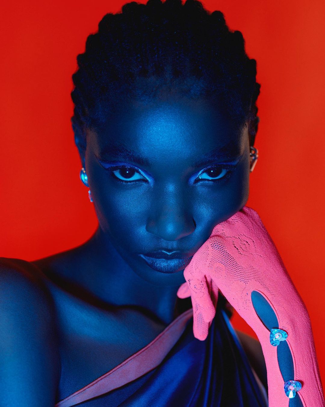 Close-up studio portrait of an African-descent adult female with braided hair and blue eyeshadow. She wears pink gloves and a draped blue top.
