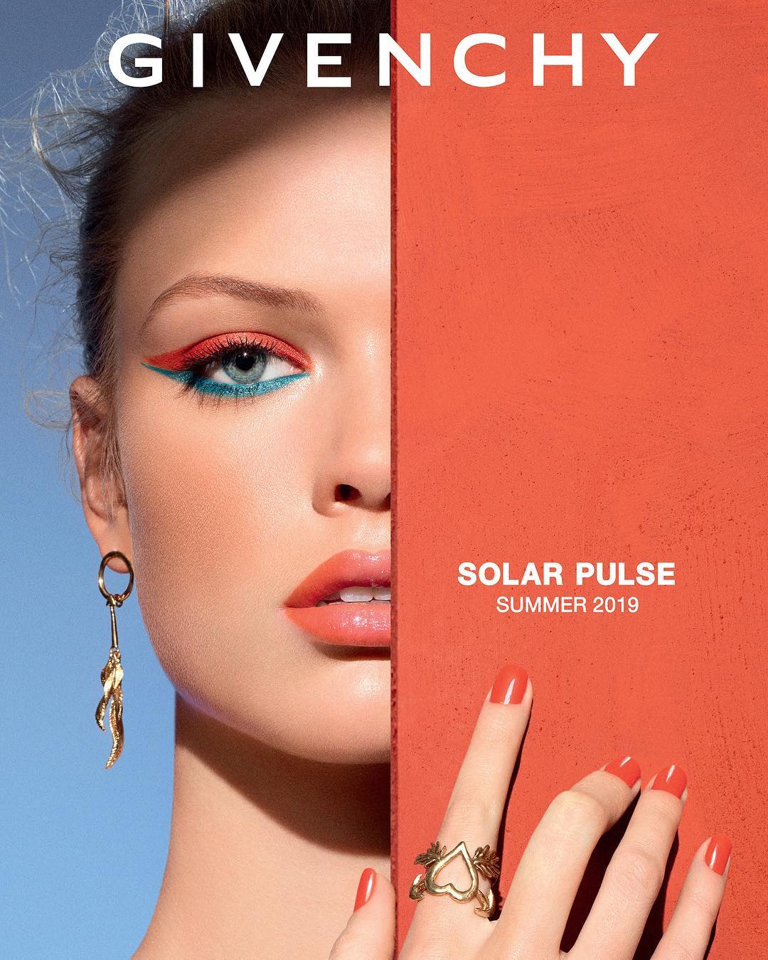 Close-up of a fair-skinned adult female model with coral lips, colorful eyeliner, and gold jewelry in a fashion ad.