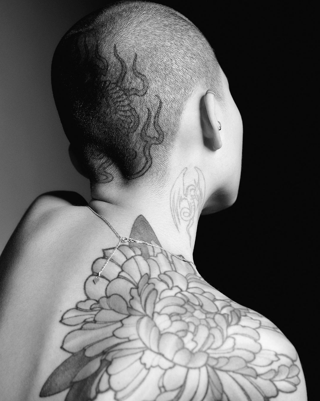 Black and white shot of a woman's upper back and head showcasing tattoos, with a partially visible silver chain necklace.