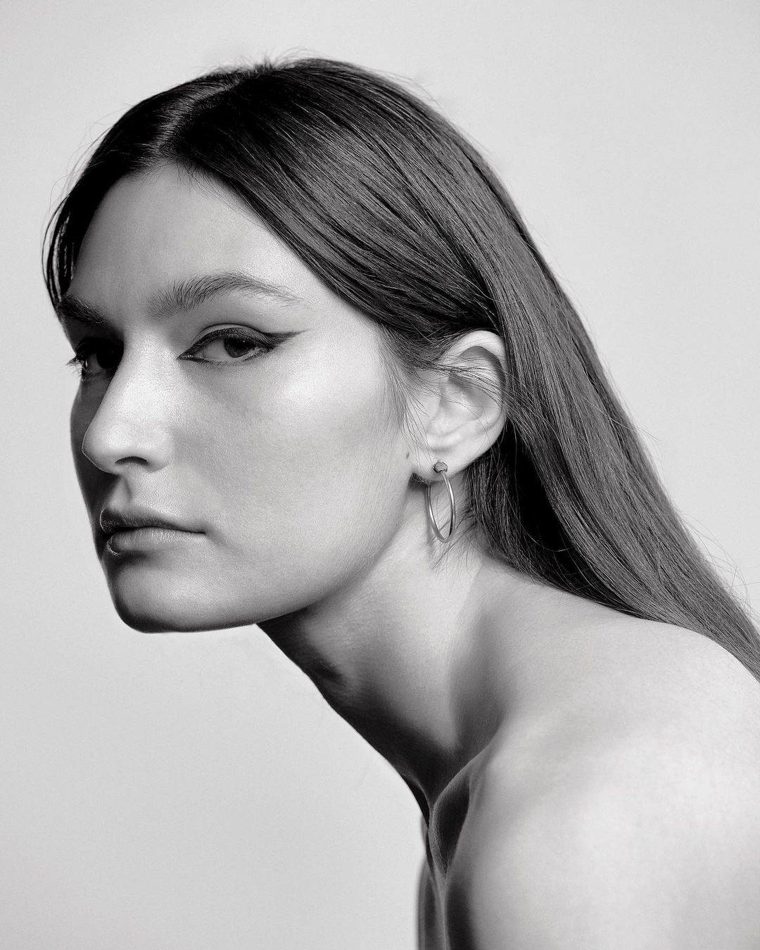 Monochrome portrait of a woman with long straight hair and winged eyeliner wearing hoop earrings against a light background