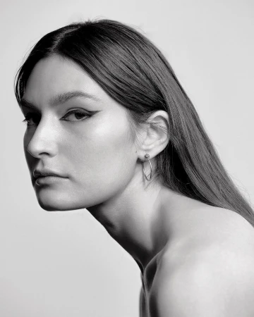 Monochrome portrait of a woman with long straight hair and winged eyeliner wearing hoop earrings against a light background
