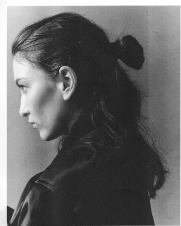 Monochrome profile portrait of a woman with dark hair tied in a bun, wearing a dark jacket against a plain background.