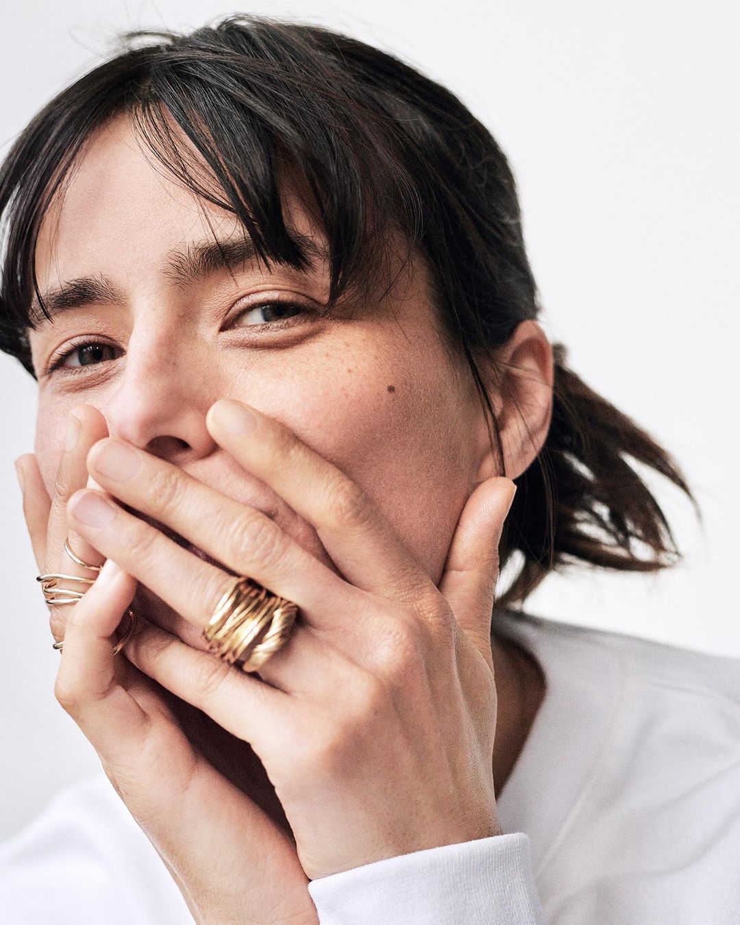 Close-up portrait of a woman with dark hair and bangs holding her hands over her mouth, wearing multiple gold rings.
