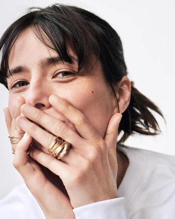 Close-up portrait of a woman with dark hair and bangs holding her hands over her mouth, wearing multiple gold rings.