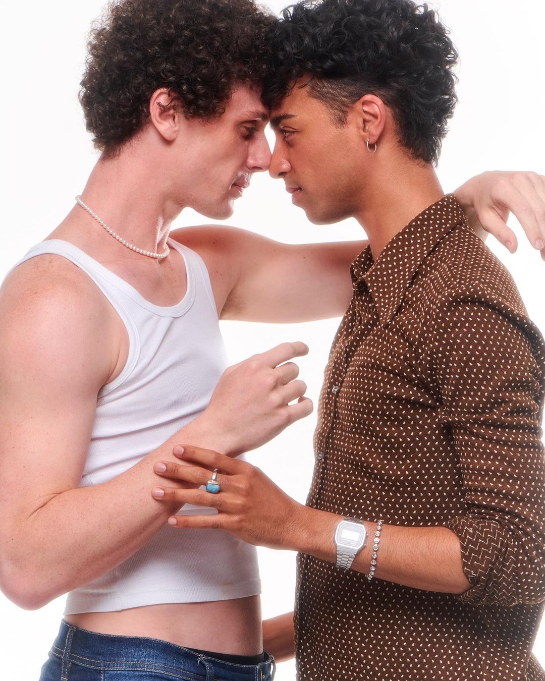 Two young men of different ethnicities stand close together in a bright, studio setting, wearing casual clothes and jewelry.