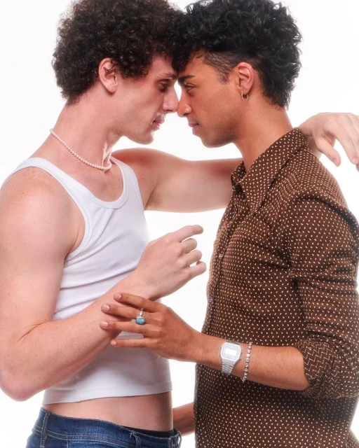 Two young men of different ethnicities stand close together in a bright, studio setting, wearing casual clothes and jewelry.