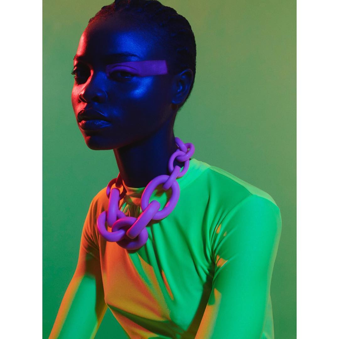 Striking studio portrait of a dark-skinned woman wearing a bright pink statement chain necklace, neon top, and vivid neon makeup.