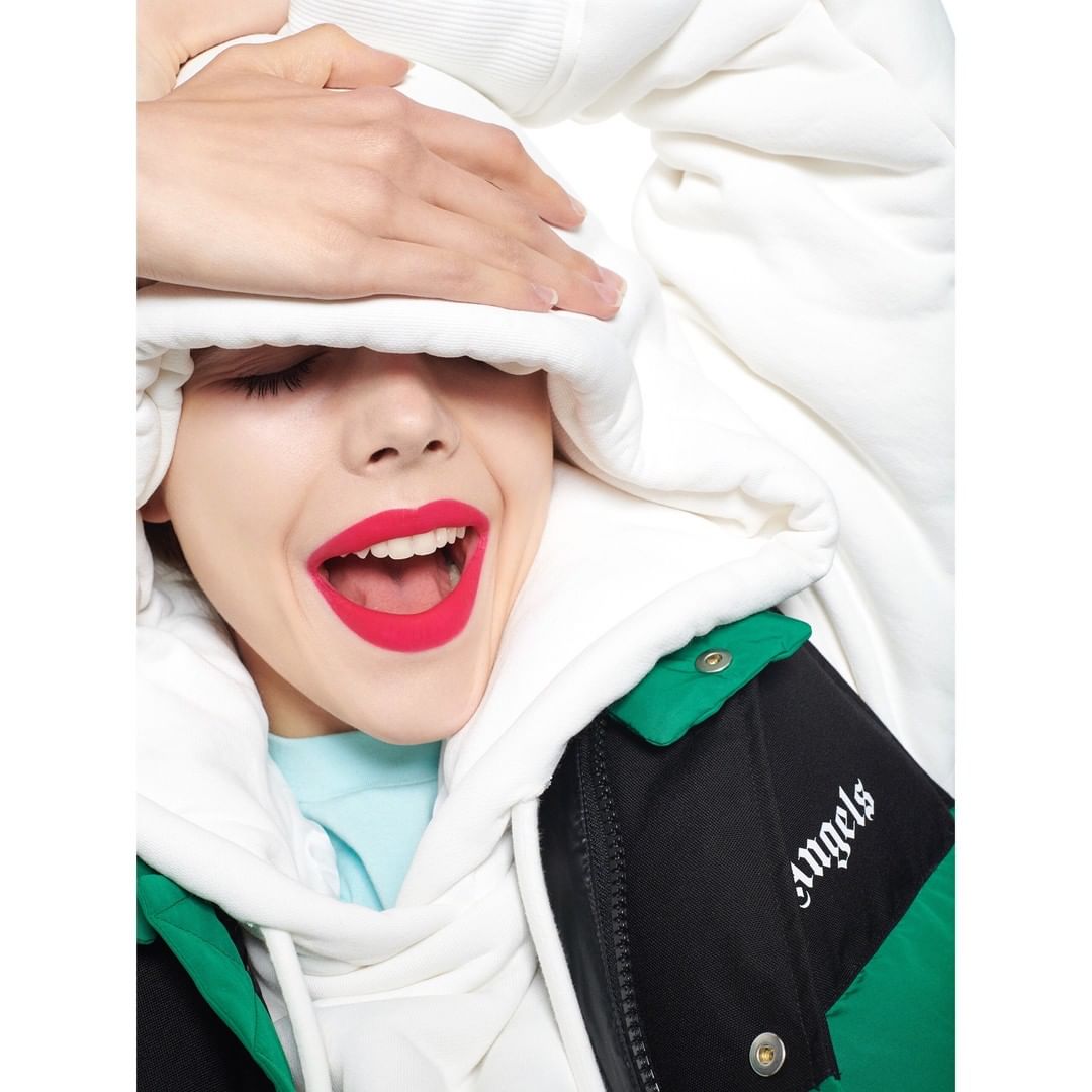 A young adult woman with pink lipstick, wearing a white hooded sweatshirt and jacket, is seen indoors with a hand covering her face.