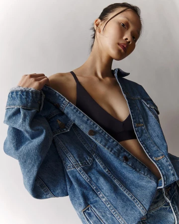 Portrait of a woman wearing a blue denim jacket slipping off her shoulder, revealing a dark bralette against a plain white background.