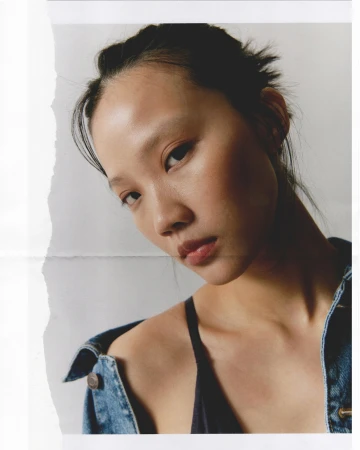 Close-up portrait of a woman wearing a denim jacket against a plain background with a torn paper overlay effect on the left edge.