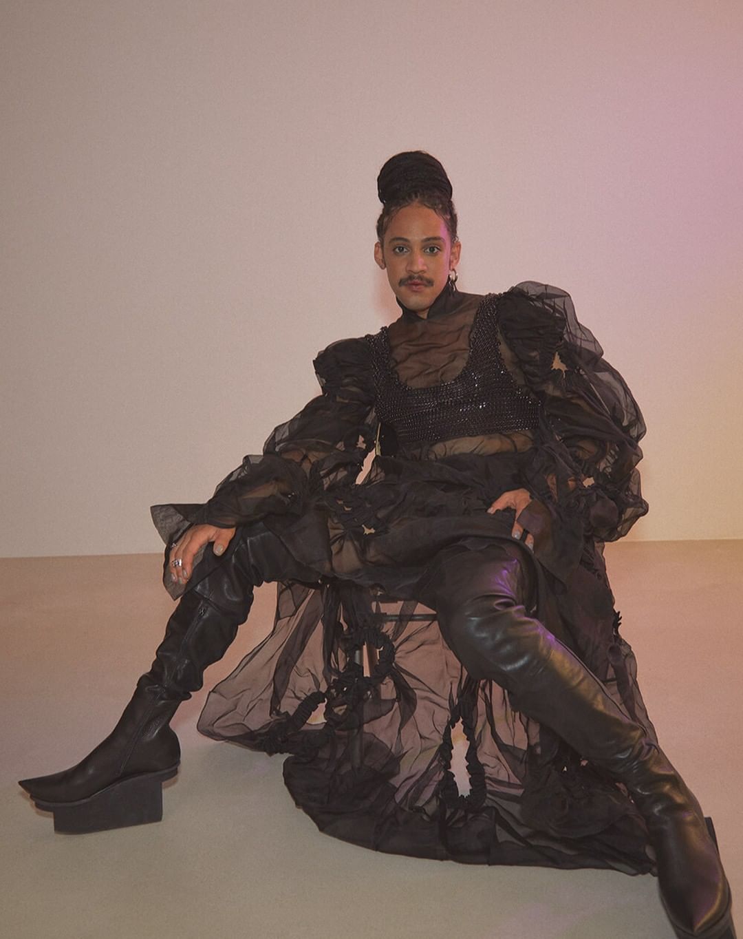 Person in Avant-Garde black outfit and chunky heel boots, posing against a solid backdrop with soft lighting and artistic styling.