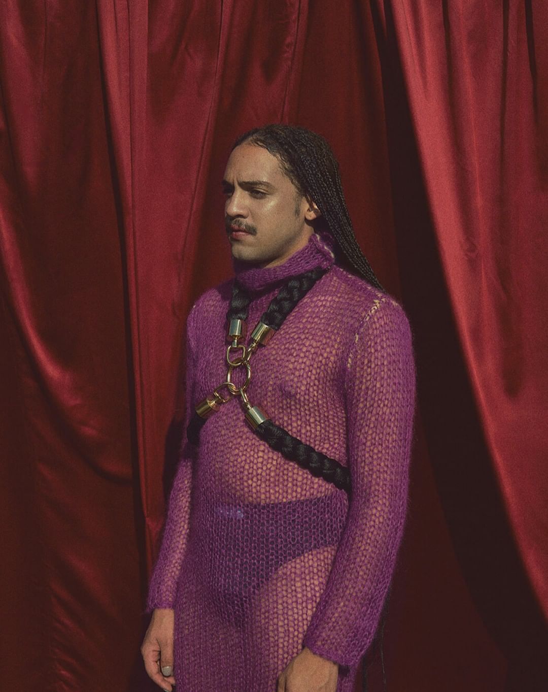 Person with long braids and a sheer purple outfit stands in front of red curtains in a studio environment, illuminated by soft light.