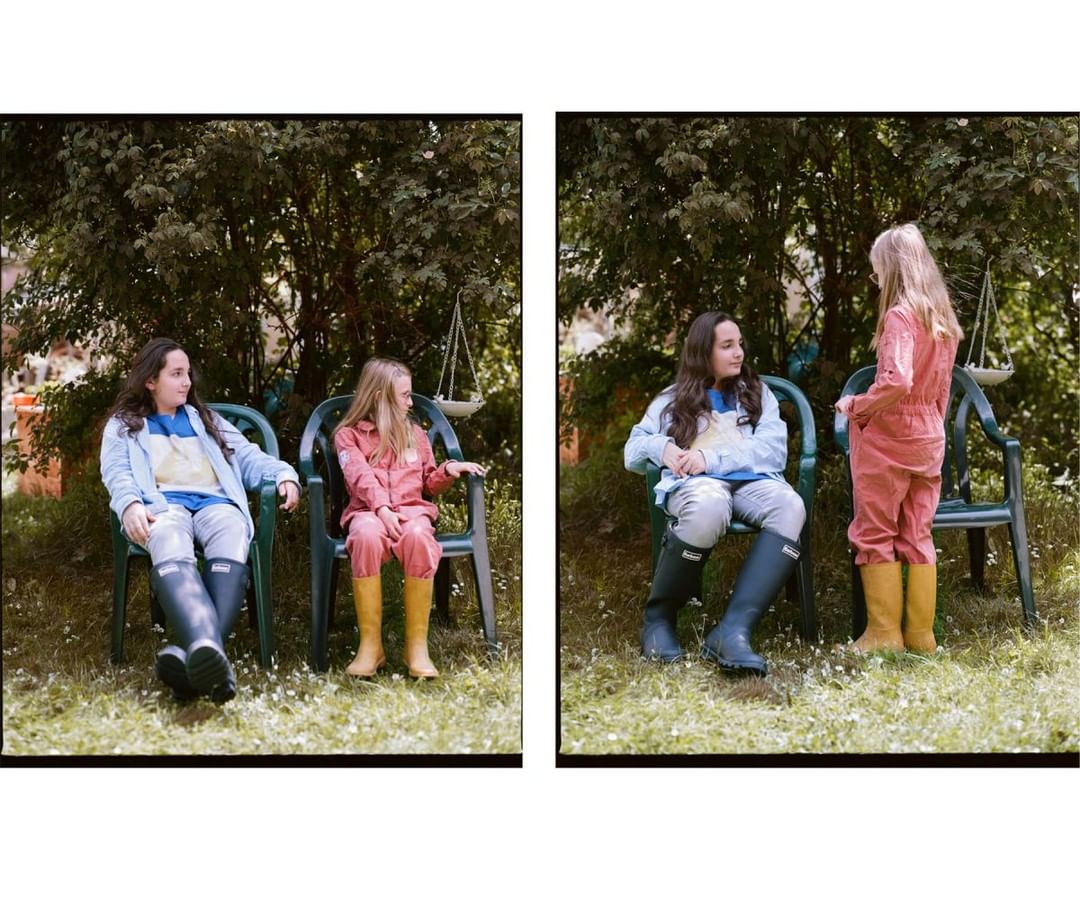 Two young girls in outdoor setting, seated on green plastic chairs, wearing casual clothing and rain boots, enjoying a natural backdrop.