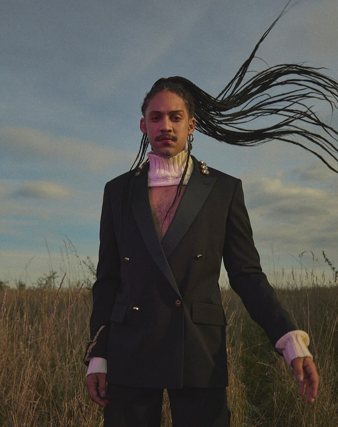 Portrait of a Black man with long braids wearing a black blazer outdoors in a field under a cloudy sky, fashion editorial style.