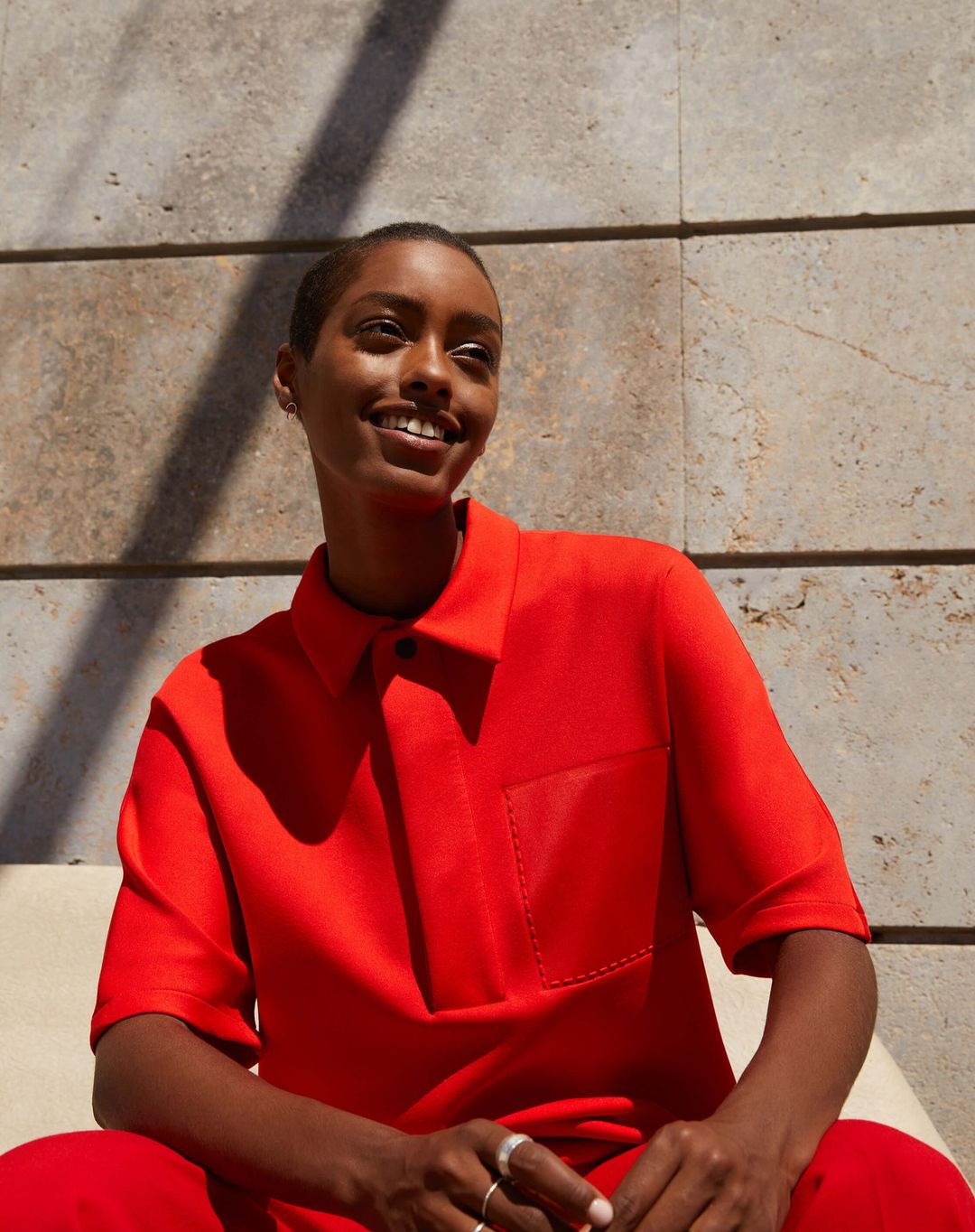 Smiling Black woman in a red polo shirt sits outdoors with a concrete wall background and strong sunlight casting a shadow on the wall.