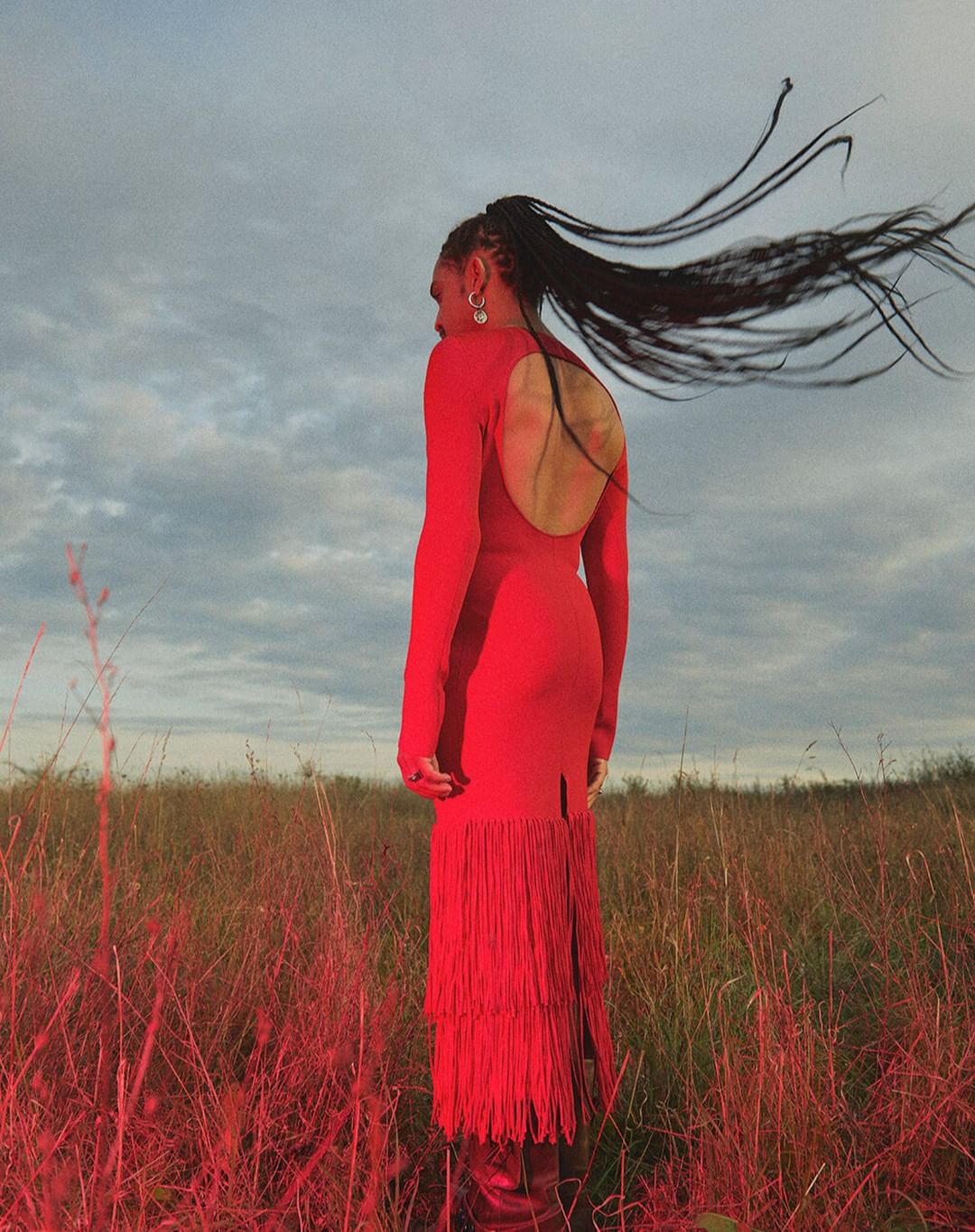 Adult female in a flowing red dress standing in a field with her hair blowing. Her back is exposed and she is looking to the side.