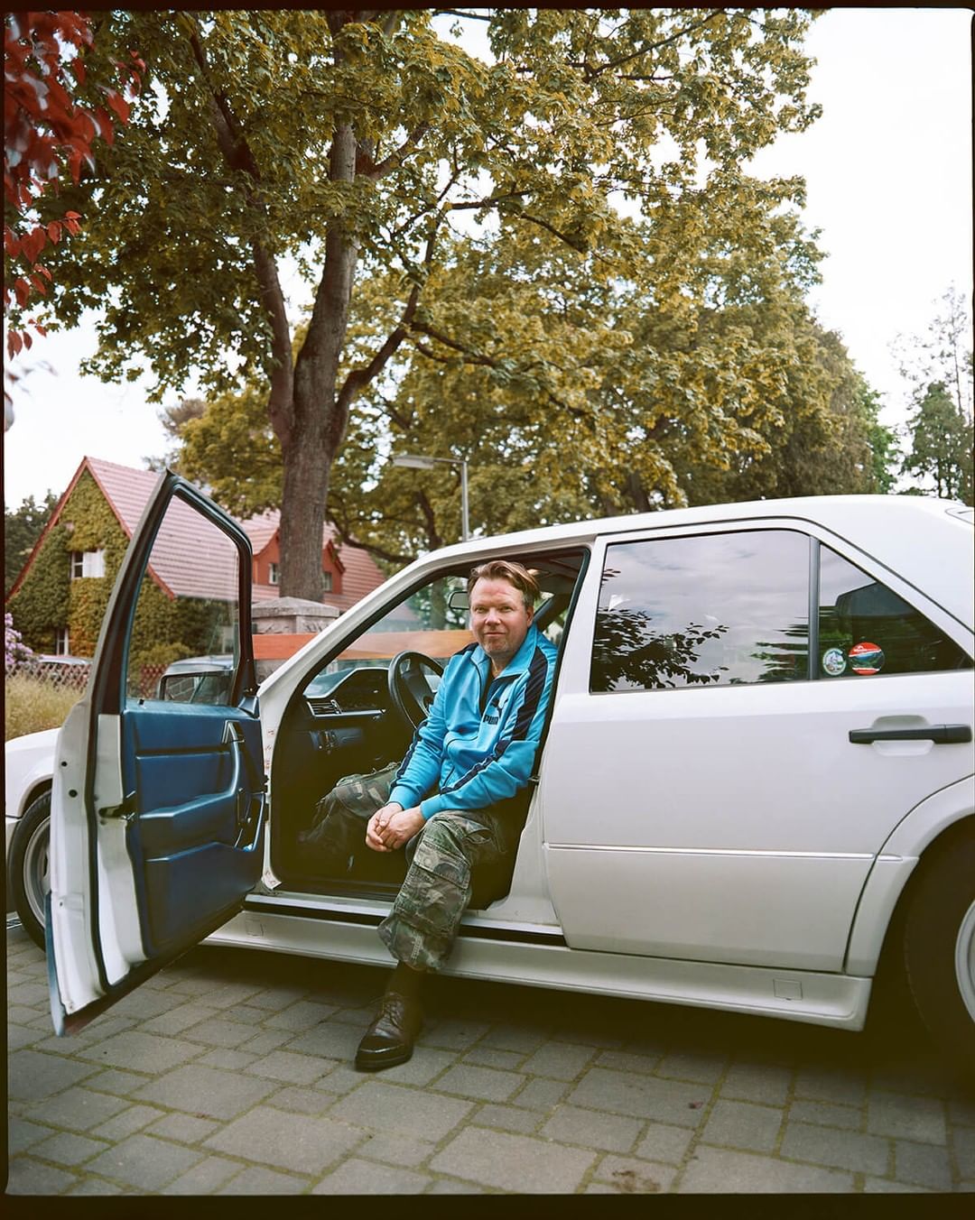 Adult male in blue jacket and camo pants sitting in the driver's seat of a white car parked outdoors on a paved surface surrounded by trees.