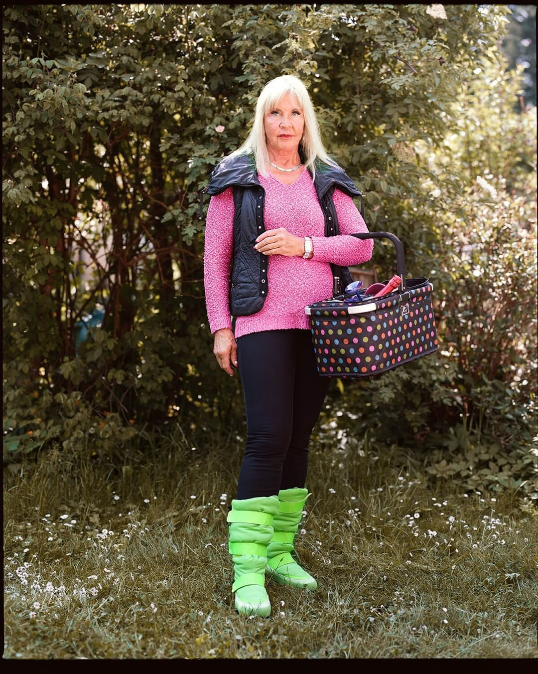 Full shot of a fair-skinned adult woman standing on a lawn in a pink sweater, black vest, and green boots, holding a polka dot basket.