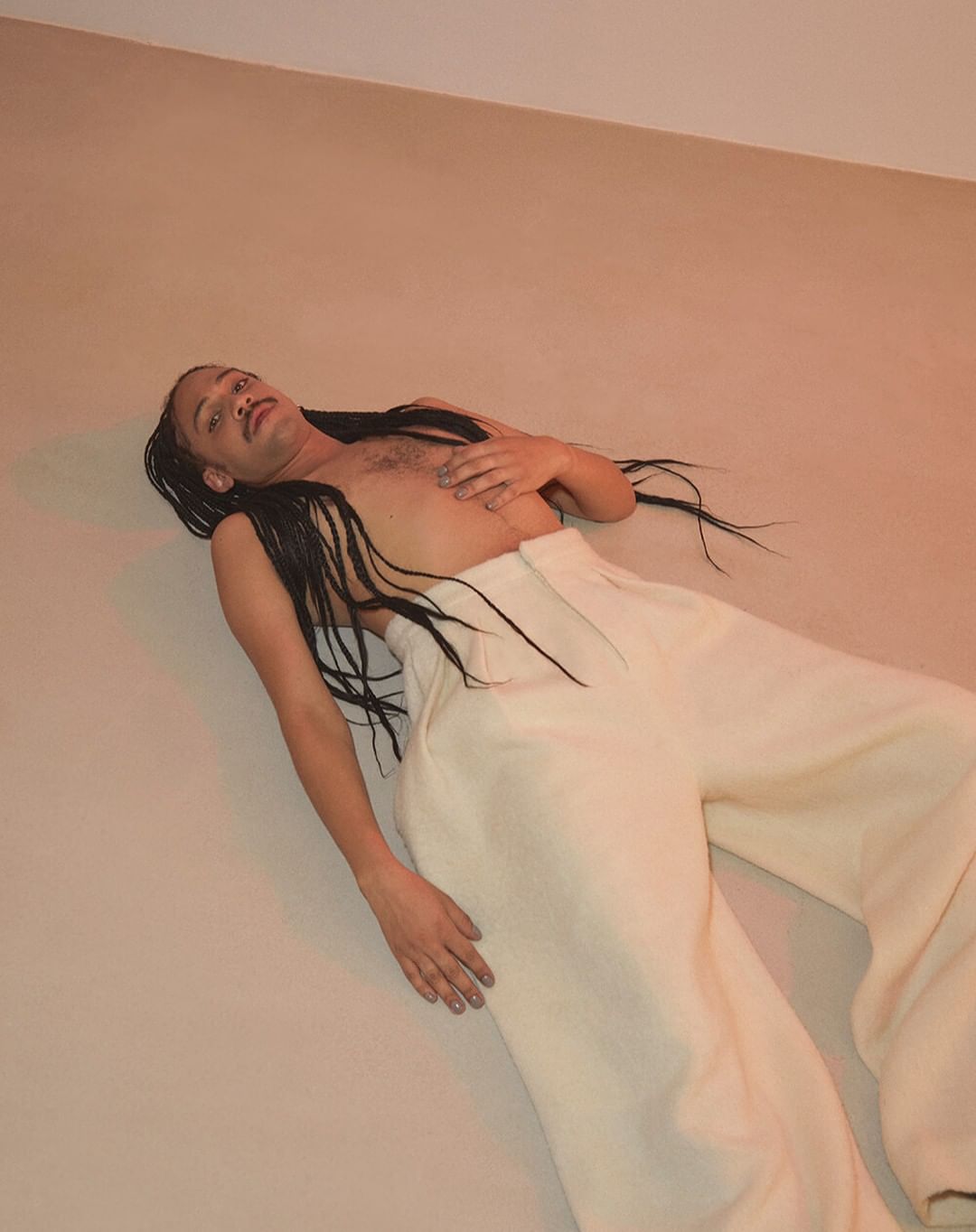 Adult male with long braids poses against a peach background, wearing white pants and shirtless in an indoor environment.